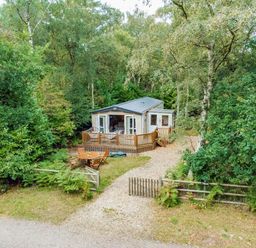 a holiday lodge surrounded by trees with a gravel path and outdoor seating at Kelling Heath Holiday Park - Holiday Accommodation 18664 in Holt