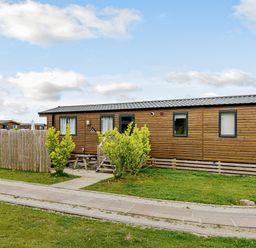 A wooden cabin with a fence and bushes in an outdoor area at Alder Waterside Spa in Carlton Miniott near Thirsk