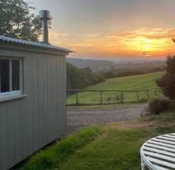 A hut with a chimney next to grass and a sunset view at Cosy Shepherd’s Hut Longhope