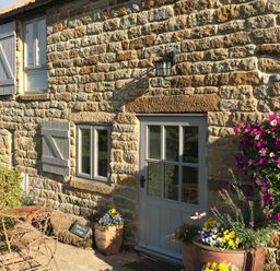 A stone cottage exterior with grey windows and door a small metal table and chairs and potted flowers at Honey Bee Cottage in Staintondale