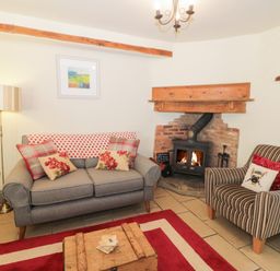 A living room with a grey sofa with cushions and a striped armchair near a brick fireplace with a burning fire at Honey Bee Cottage in Staintondale