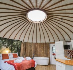 A round room with a bed dressed with orange pillows and a blanket a bathtub next to wooden paneled walls and a kitchen area with a sink and cabinets under a circular ceiling with wooden beams Lakeview Yurt Beckford near Tewkesbury