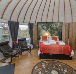 A bedroom with a bed with red bedding two black chairs windows with dark curtains and a bathtub against wooden walls at Lakeview Yurt in Beckford near Tewkesbury