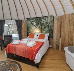 A bedroom with a double bed white and orange linens two black chairs wooden walls and a white bathtub with a towel at Lakeview Yurt in Beckford near Tewkesbury