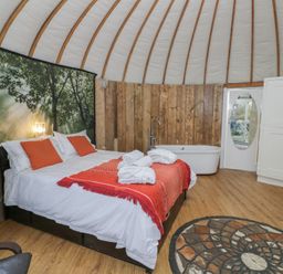 A bedroom with a double bed with orange pillows and blanket a bathtub a circular patterned rug and wooden walls at Lakeview Yurt Beckford near Tewkesbury
