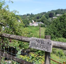 A sign on a fence indicating 'MILL BARN' with hills and trees in the background at The Mill Barn in Lustleigh