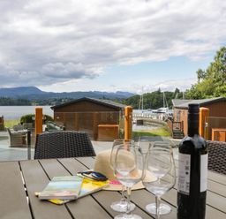An outdoor area with a table, glasses, and a view of water and cabins at 19 Pony Meadow White Cross Bay near Troutbeck Bridge