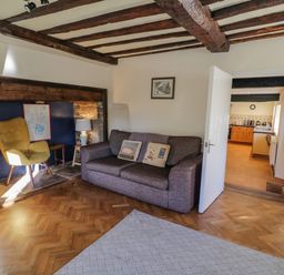 A living room with a gray sofa, a yellow chair, a side table with a lamp, wooden beams on the ceiling, and a kitchen visible through an open door at Little Cowarne Court in Little Cowarne near Bromyard