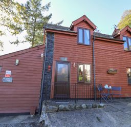 The exterior of a red wooden cottage with a door labeled cedar and two blue chairs with a table outside at Cedar Cottage in Llanidloes