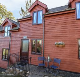 Exterior of a red wooden cottage with windows two chairs and table outside at Beech Cottage in Llanidloes