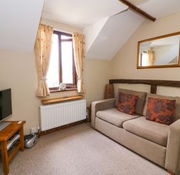 A small living room with a beige sofa with two patterned cushions a wall mirror a window with curtains a radiator a wooden side table with a TV and a stereo unit at Oak Cottage in Llanidloes