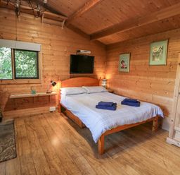 A bedroom with a wooden bed with blue towels on it a wooden wardrobe a window a wall-mounted television and a wood-burning stove at The Log Cabin in Oban
