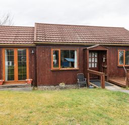A wooden house with a tiled roof and glass door next to windows and outdoor chairs on grass at Glen Dessary in Corpach near Fort William