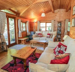 A wooden living room with two beige sofas red and floral cushions wooden tables and floral curtains at Striding Edge in White Cross Bay near Troutbeck Bridge