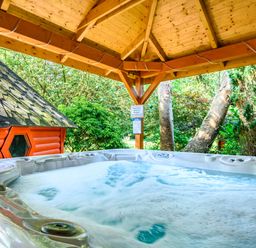 A bubbling hot tub under a wooden gazebo with trees and a small wooden cabin nearby at Eden House in Bardsea