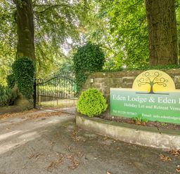 A gated entrance with trees and stone walls showing a sign for Eden Lodge and Eden House in Bardsea