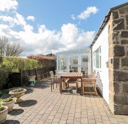 An outdoor patio area with a wooden table and chairs next to a house with a stone wall and glass doors at Lyndhurst Cottage in Beadnell