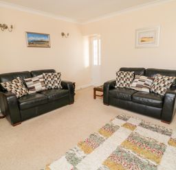 A living room with two black leather sofas with cushions a small wooden side table with a lamp a patterned rug and framed pictures on the walls at Lyndhurst Cottage in Beadnell