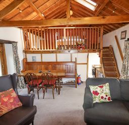 A living area with wooden dining table and chairs, a dark armchair, a sofa with floral cushions, wooden staircase and railing at The Shippon in Llechwedd The Conwy Valley