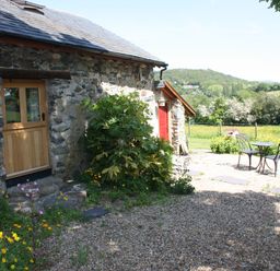 A stone cottage with a wooden door and a red door next to a garden with flowers and a metal table and chairs set outside at The Shippon in Llechwedd The Conwy Valley