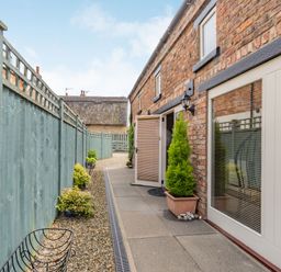 A narrow outdoor pathway with gravel and plants along a brick wall next to a green wooden fence at Upstairs Downstairs Cottage in Snainton near Scarborough