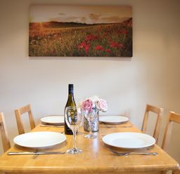 A dining table set with white plates wine glasses cutlery and a bottle of wine under a wall painting of a field at Yew Tree Cottage in Little Malvern near Malvern