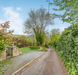 A narrow country lane with green hedges and a gravel driveway with a stone wall and gate at Hillside Cottage in Peasedown St John near Bath