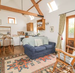 A room with wooden beams a blue sofa a rocking chair a patterned rug a dining table and a small kitchen at High Park Barn in Uffculme Blackdown Hills