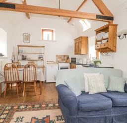 A living area with a blue sofa and pillows a dining table with wooden chairs and a kitchen with wooden cabinets and shelves at High Park Barn in Uffculme Blackdown Hills