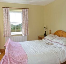 A bedroom with a wooden bed and bedside table a standing mirror and window with floral curtains at The Bay in Caherdaniel County Kerry
