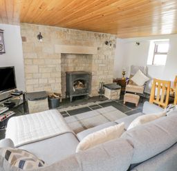 A living room with a stone fireplace a wood stove a gray sofa a TV on a glass stand and a wooden dining table with chairs at Mary Kates Cottage in Narin County Donegal