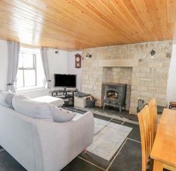 A living room with a gray sofa a wood stove a TV and a wooden dining table at Mary Kates Cottage in Narin County Donegal