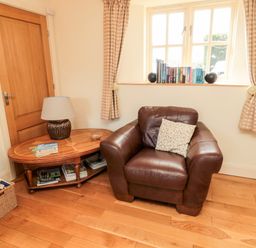 A room corner with a brown leather armchair a wooden side table with a lamp books on it and a window with books and curtains at The Granary Longframlington near Alnwick
