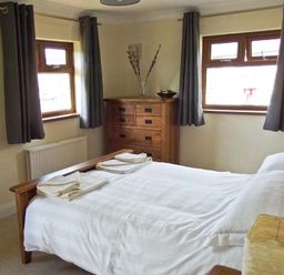 A bedroom with a wooden bed white bedding folded towels a wooden chest of drawers two windows with dark curtains and a small lamp on bedside table at Hartam House in Falstone near Kielder and Bellingham