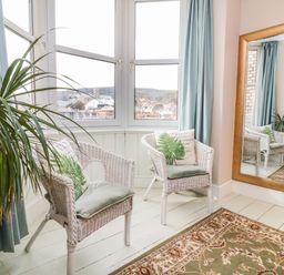A room with two white wicker chairs with cushions near a large window and a tall mirror at Penryn in Ilfracombe