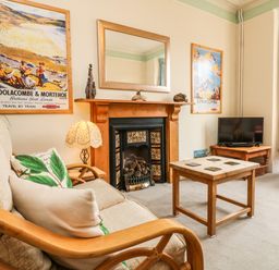 A living room with a beige armchair and pillows a wooden coffee table a fireplace with a mirror above and a small television on a wooden stand at Penryn in Ilfracombe