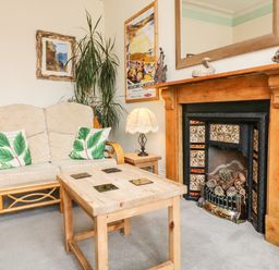 A sitting area with a wooden framed sofa with cushions a wooden coffee table a side table with a lamp a fireplace with decorative tiles and pinecones a tall plant and framed pictures on the walls at Penryn in Ilfracombe