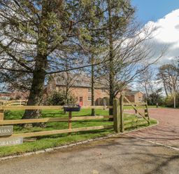A brick house behind a wooden fence and gate with trees and a gravel driveway at The Red Barn in Longframlington near Alnwick