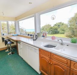A kitchen with a countertop sink and two bar stools by a corner wooden table near large windows showing a garden and sea view at Morfa Isaf Cottage in Llangrannog