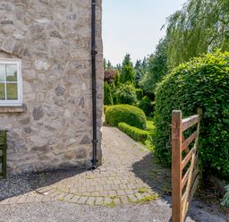 An outdoor garden area with a stone wall house a gate and a paved path surrounded by trimmed bushes at Distyll Llanfwrog near Ruthin