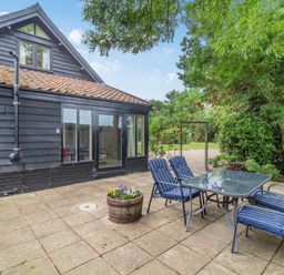 An outdoor patio with a glass table and four chairs with blue cushions outside a black wooden house at Ducksfoot Barn Bush Green near Pulham Market