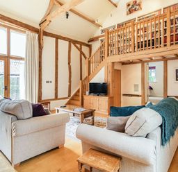 A living room with two beige sofas a wooden coffee table a TV on a wooden cabinet stairs leading to a bookshelf balcony and large windows at Ducksfoot Barn in Bush Green near Pulham Market