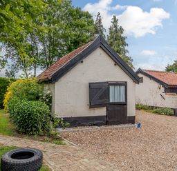 A small white brick building with a tiled roof and black door surrounded by gravel and greenery at Garden Cottage in Bush Green near Pulham Market