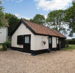 A small white cottage with black trim and a tiled roof surrounded by trees and lawn at Garden Cottage in Bush Green near Pulham Market