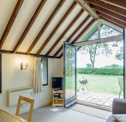 A room with wooden beams a dining table chairs a TV stand a sofa and an open glass door leading to a garden at Garden Cottage Bush Green near Pulham Market