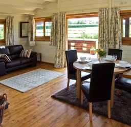 A dining table with four chairs and a vase of flowers near a black sofa with cushions and windows at Cropvale Lodge in Cropthorne near Pershore