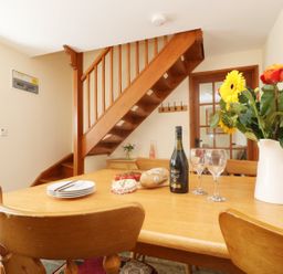 A dining room with wooden table and chairs flowers wine bread and cheese on the table and wooden stairs in the background at The Old Granary in Polyphant near Launceston