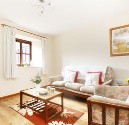 A living room with a floral sofa set a wooden coffee table with flowers and mugs a window with white curtains and a painting on the wall at The Old Granary in Polyphant near Launceston