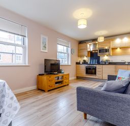 A living area with a gray sofa a wooden TV stand with a television two windows and a kitchen with wooden cabinets at Sunrise The Bay near Filey