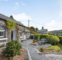 An outdoor patio with benches and a table with chairs beside a stone building at Henrhiw Bach in Penmachno